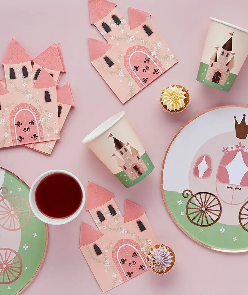Princess-themed party tableware with castle-print plates, cups, napkins, and cupcakes arranged on a pastel pink background. Princess-themed party tableware with castle-print plates, cups, napkins, and cupcakes arranged on a pastel pink background.